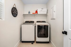 Uma lavanderia com duas máquinas de lavar, uma secadora, uma prateleira com produtos de lavagem e uma placa de lavanderia fixada na parede.