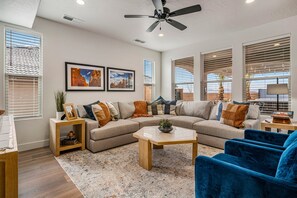 Un salon moderne avec un canapé d’angle, une table basse et un fauteuil bleu. Les murs sont ornés de tableaux encadrés, le plafond est équipé d’un ventilateur et de grandes fenêtres sont dotées de stores.