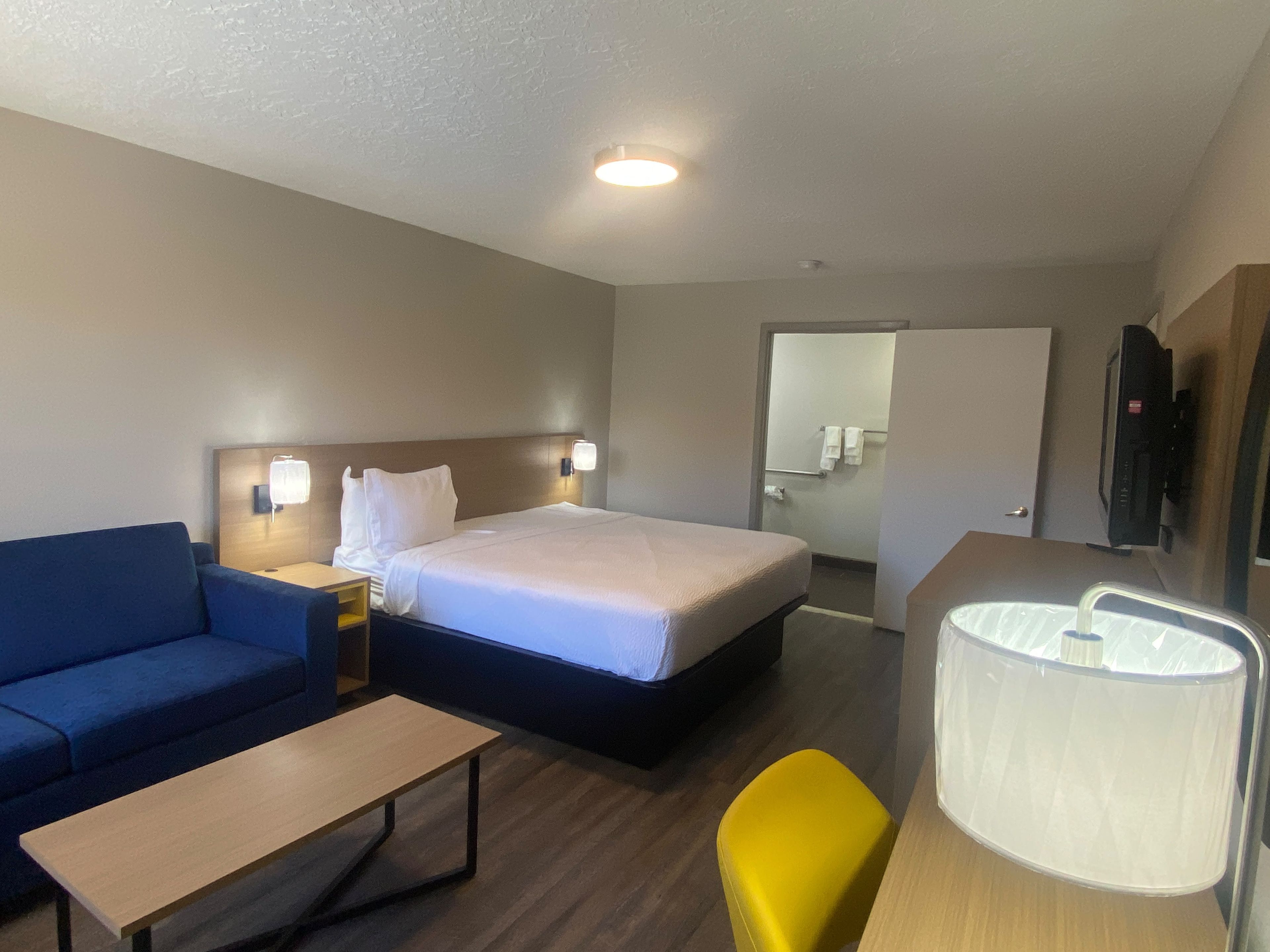 A hotel room with a bed, a sofa, a small table, a chair, a TV, and a bathroom visible through an open door.