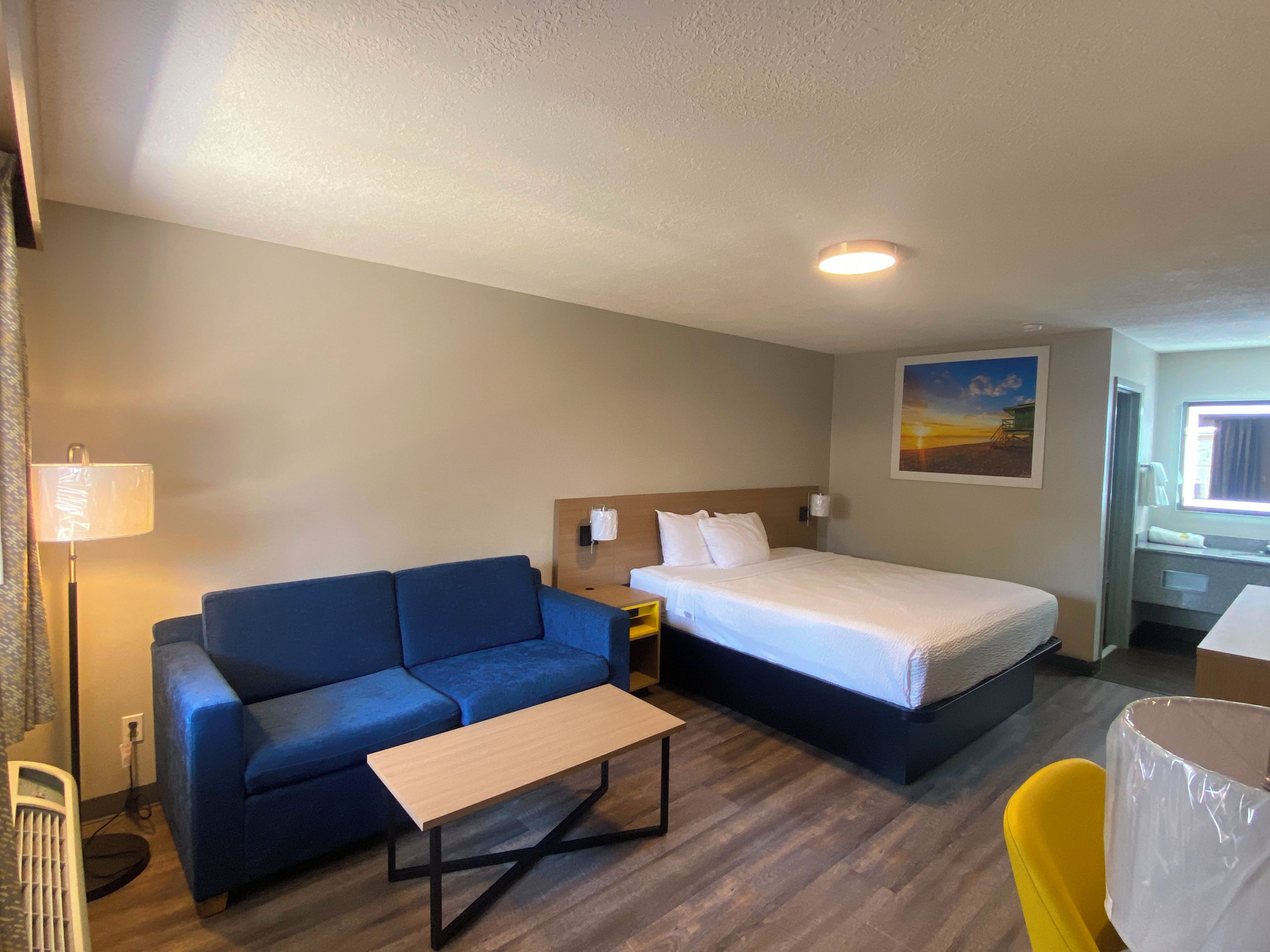 A hotel room with a bed, a sofa, a small table, a lamp, and a painting on the wall.
