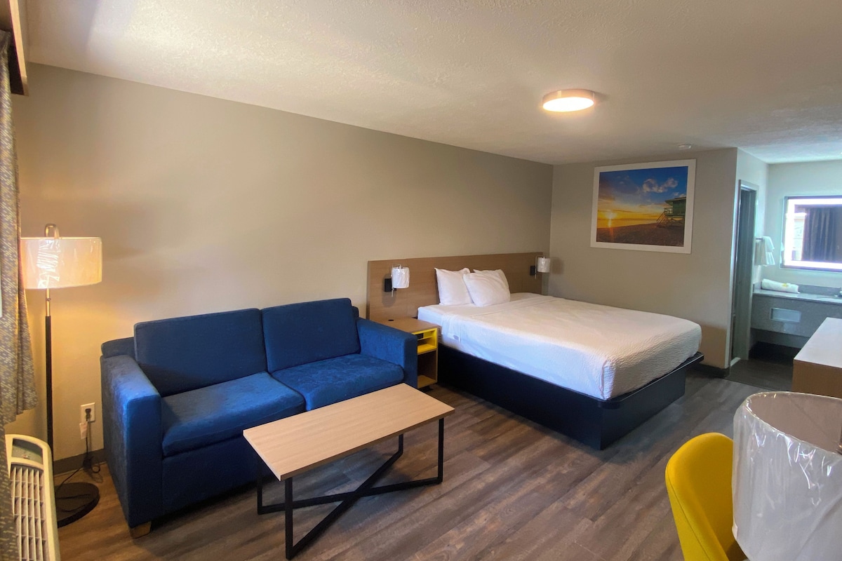 Habitación de hotel con cama, sofá, una mesita, lámpara y un cuadro en la pared.