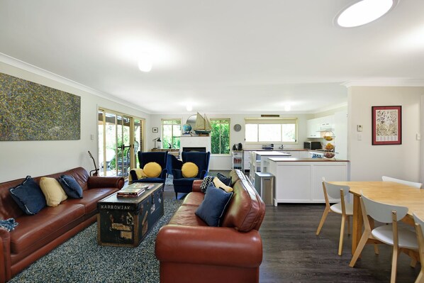 A spacious living room with a brown leather sofa, a coffee table, and a blue armchair. A kitchen with white cabinets and a dining area with a wooden table and chairs is visible in the background.