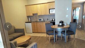 A compact kitchen with wooden cabinets, a microwave, and a small refrigerator. There is a round table with four chairs and a sofa in the living area. - One Bedroom for Icelandic Festival week (Gimli)