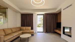 A modern living room with a beige sectional sofa, a round coffee table, a flat-screen TV mounted on the wall, and a sliding glass door leading to an outdoor area.