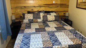 Een slaapkamer met een houten hoofdbord, een lappendeken, twee nachtkastjes met lampen en een klein plankje met decoratieve objecten.