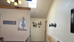 Een hal met een houten eettafel en stoelen, een 'WELCOME' bord, een decoratieve boom en een muur met een berg silhouet.