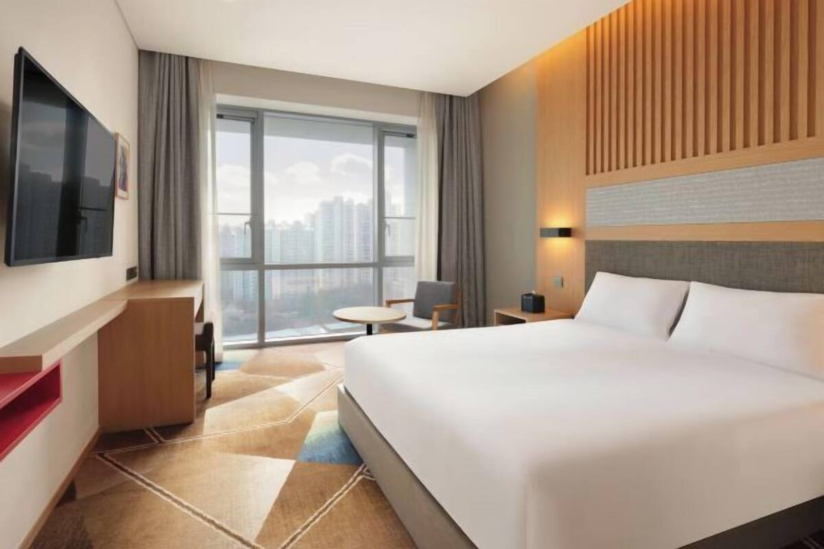 A hotel room with a large bed, a desk, a TV, a window with a city view, and a modern design.