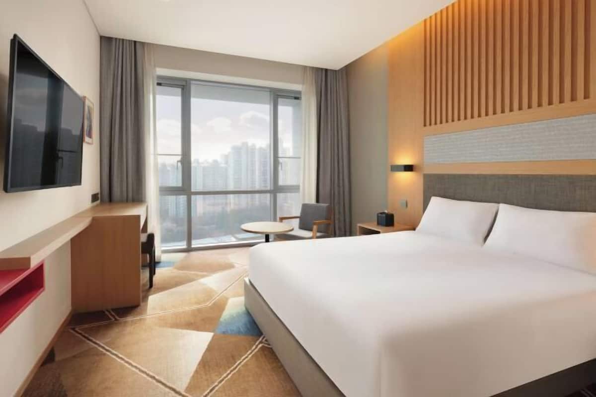 A hotel room with a large bed, a desk, a TV, a window with a city view, and a modern design.