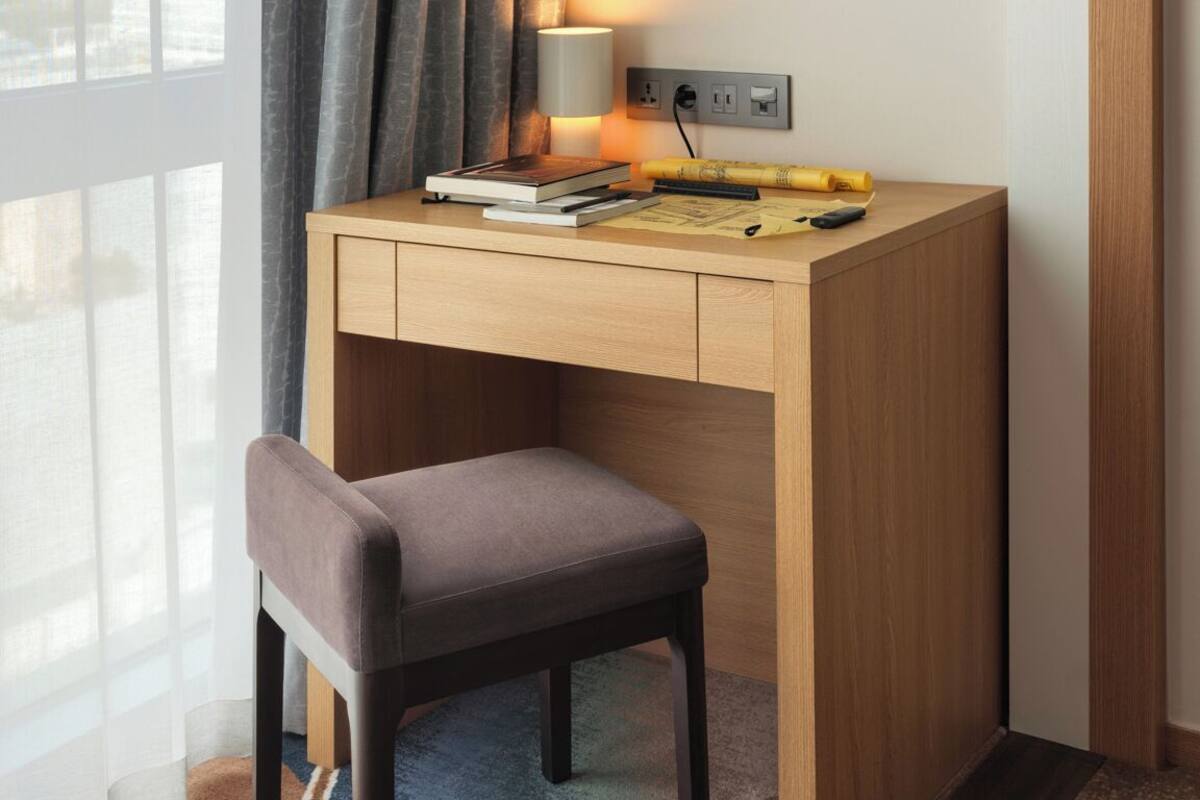 A hotel room with a wooden desk, a lamp, a chair, and a framed artwork on the wall.