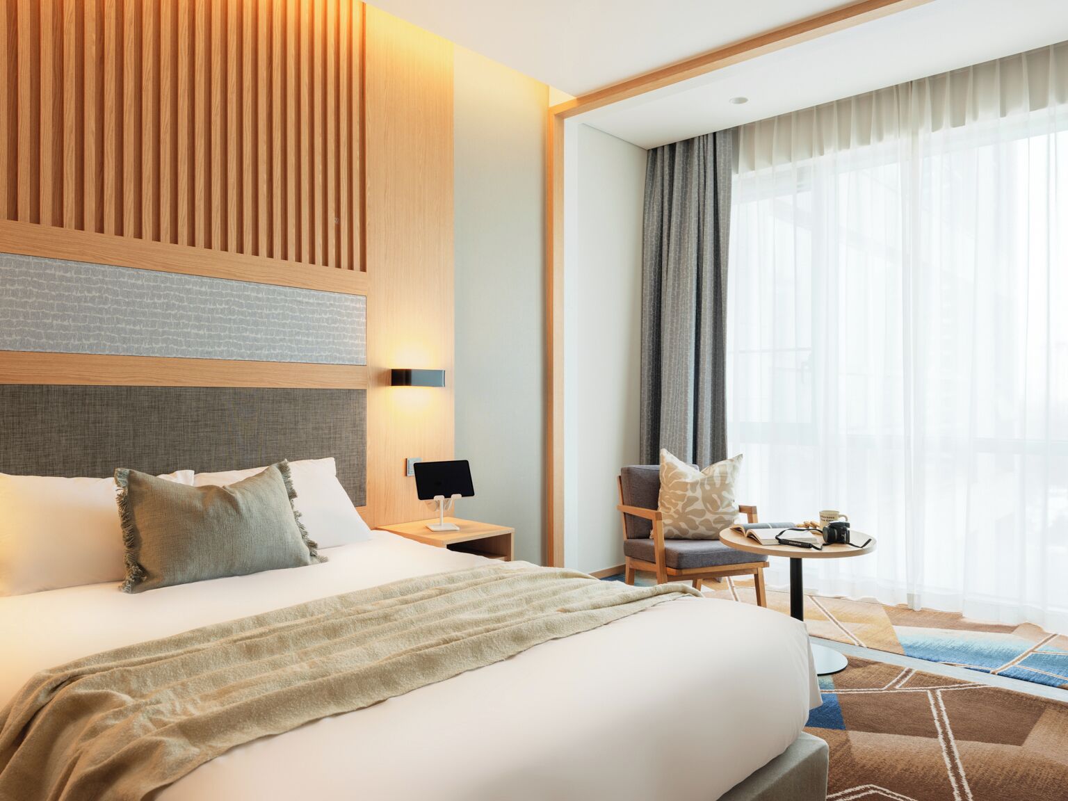 A hotel room with a large bed, a bedside table with a lamp, a chair, a small table with a cup and a book, and a large window with curtains.