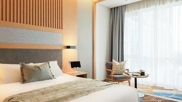 A hotel room with a large bed, a bedside table with a lamp, a chair, a small table with a cup and a book, and a large window with curtains.