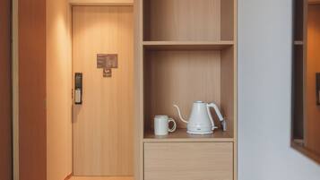 A hotel room with a wooden door, a small cabinet with a kettle and a mug, and a wall-mounted thermostat.