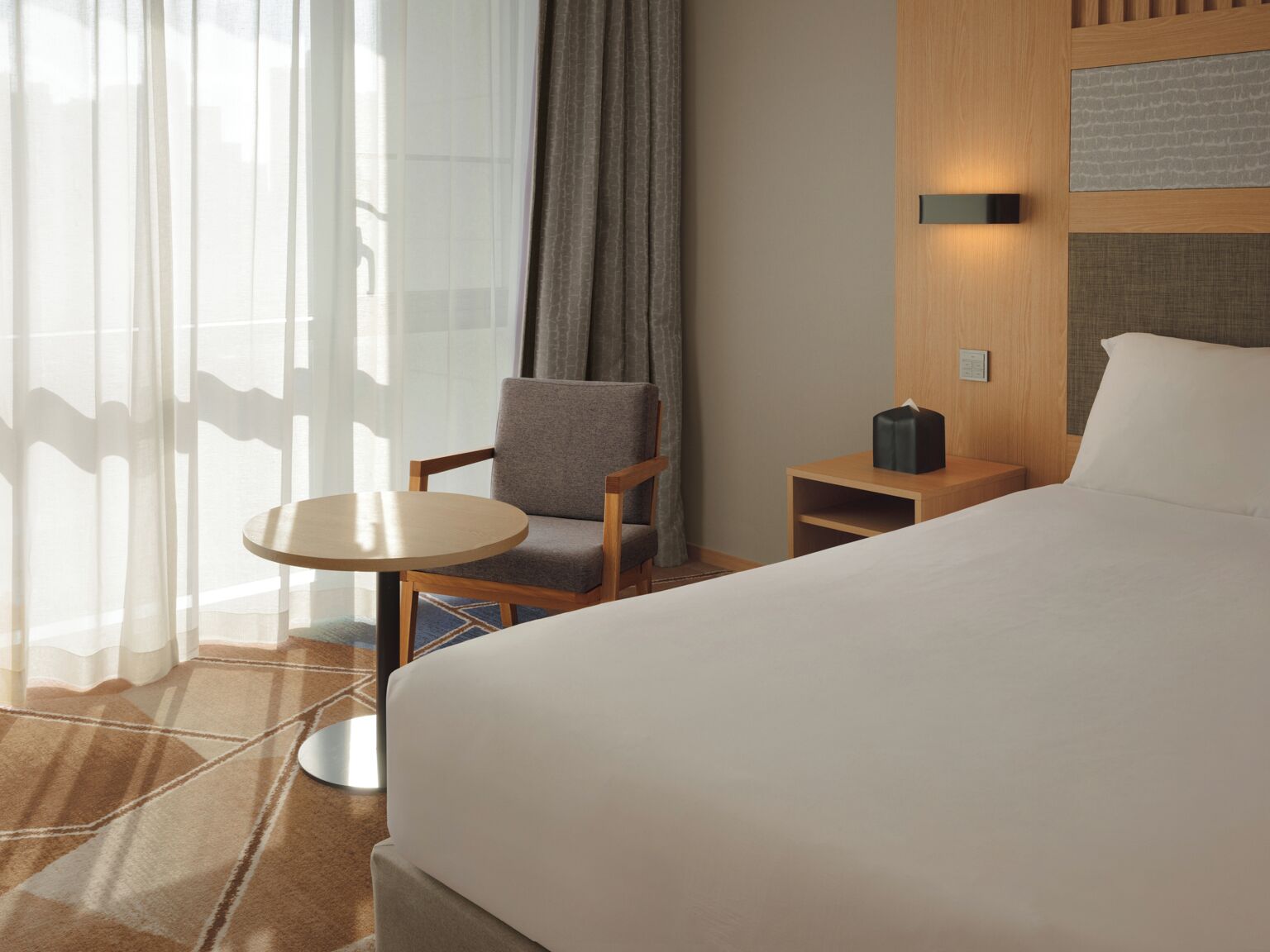 Habitación de hotel con una cama grande, una mesita redonda pequeña, una silla, una mesita de noche y una ventana con cortinas translúcidas.