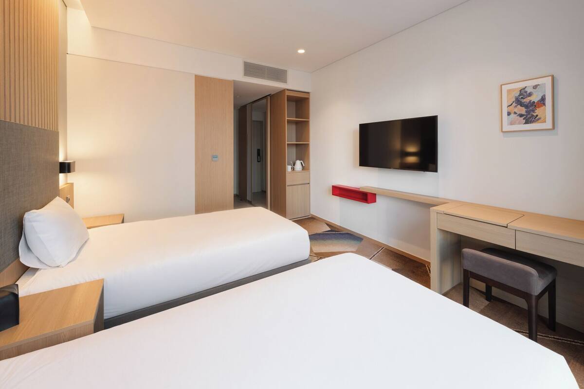 A hotel room with two beds, a desk, a chair, a TV, and a painting on the wall.