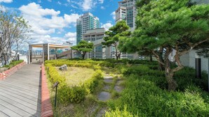 Un edificio moderno con fachada de vidrio, terraza de madera y un jardín cuidado con diversas plantas y un árbol.