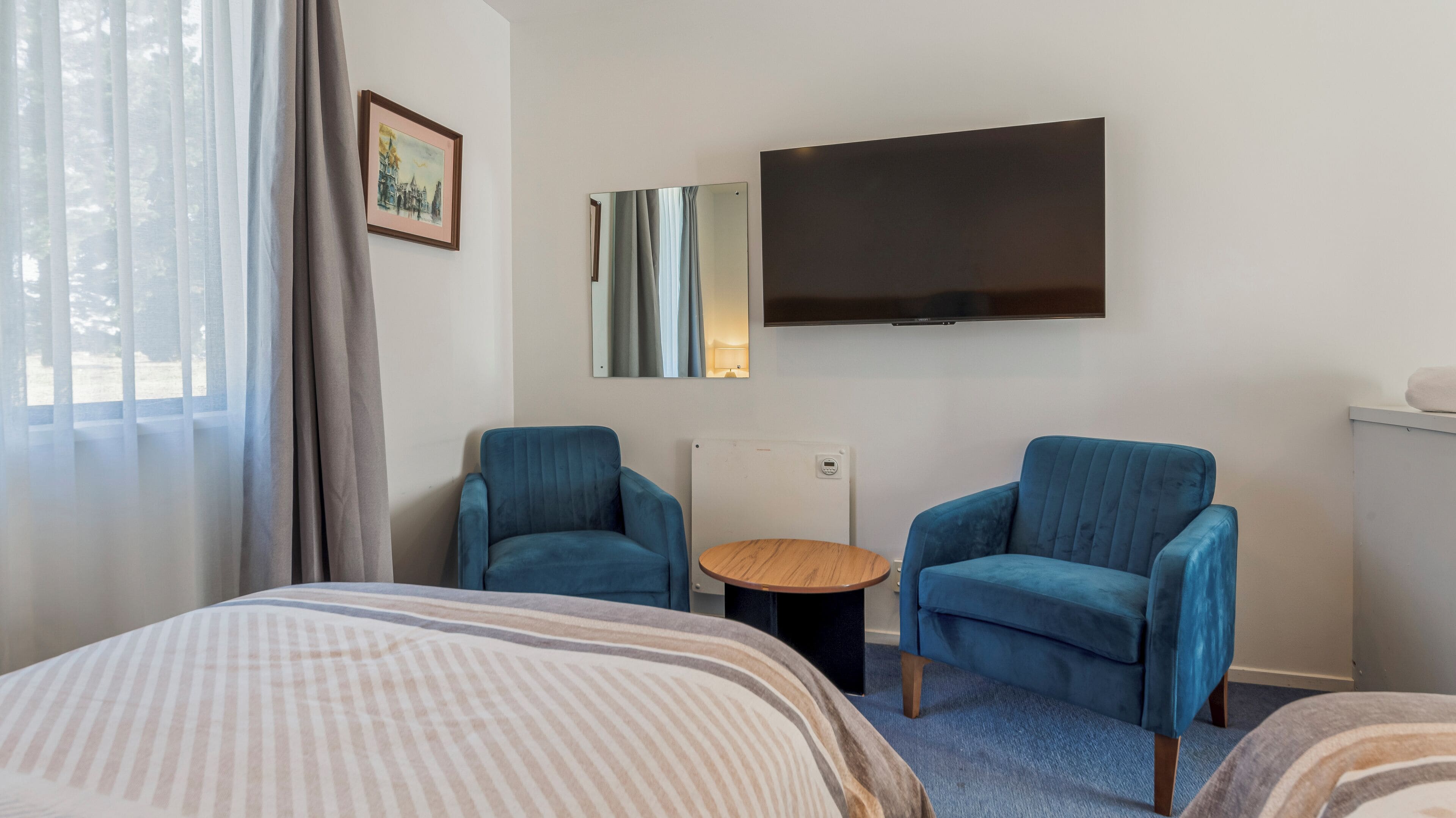 A hotel room with two blue armchairs, a bed with striped bedding, a round wooden table, a flat-screen TV mounted on the wall, and a window with sheer curtains.