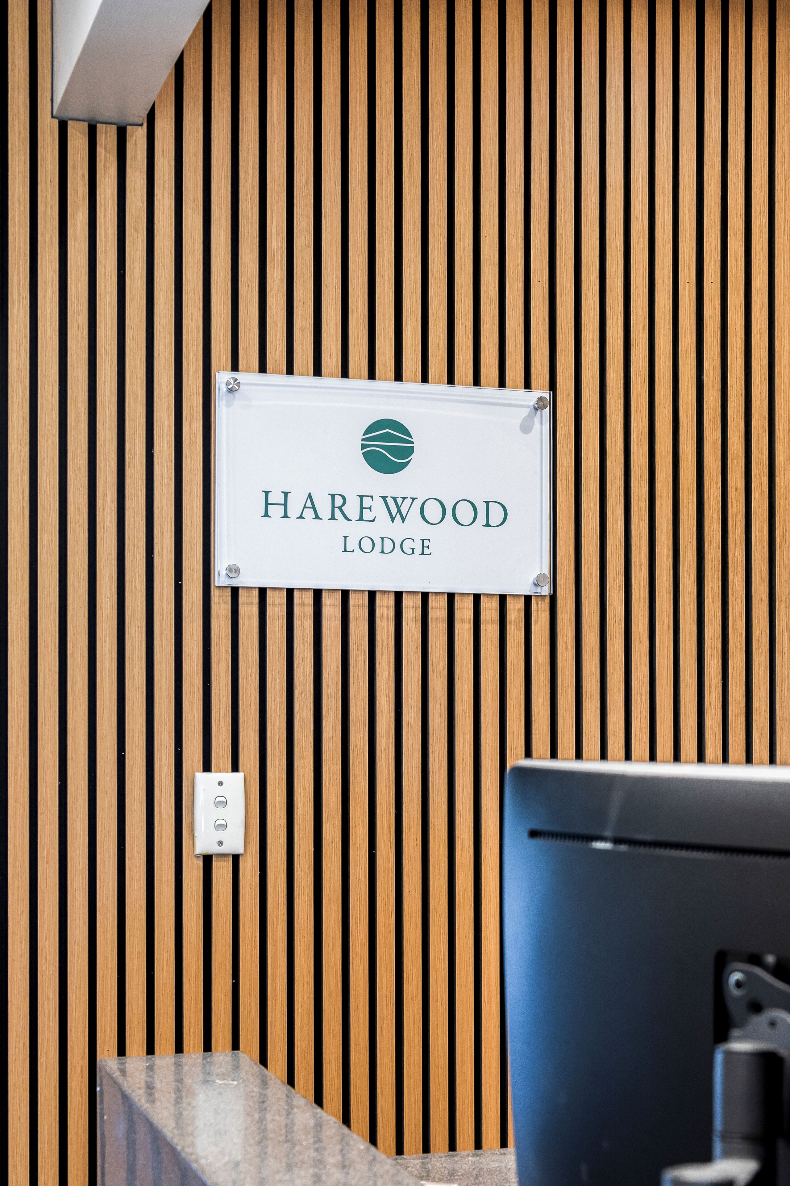 A reception desk with a sign reading "Harewood Lodge" against a wooden slat wall.