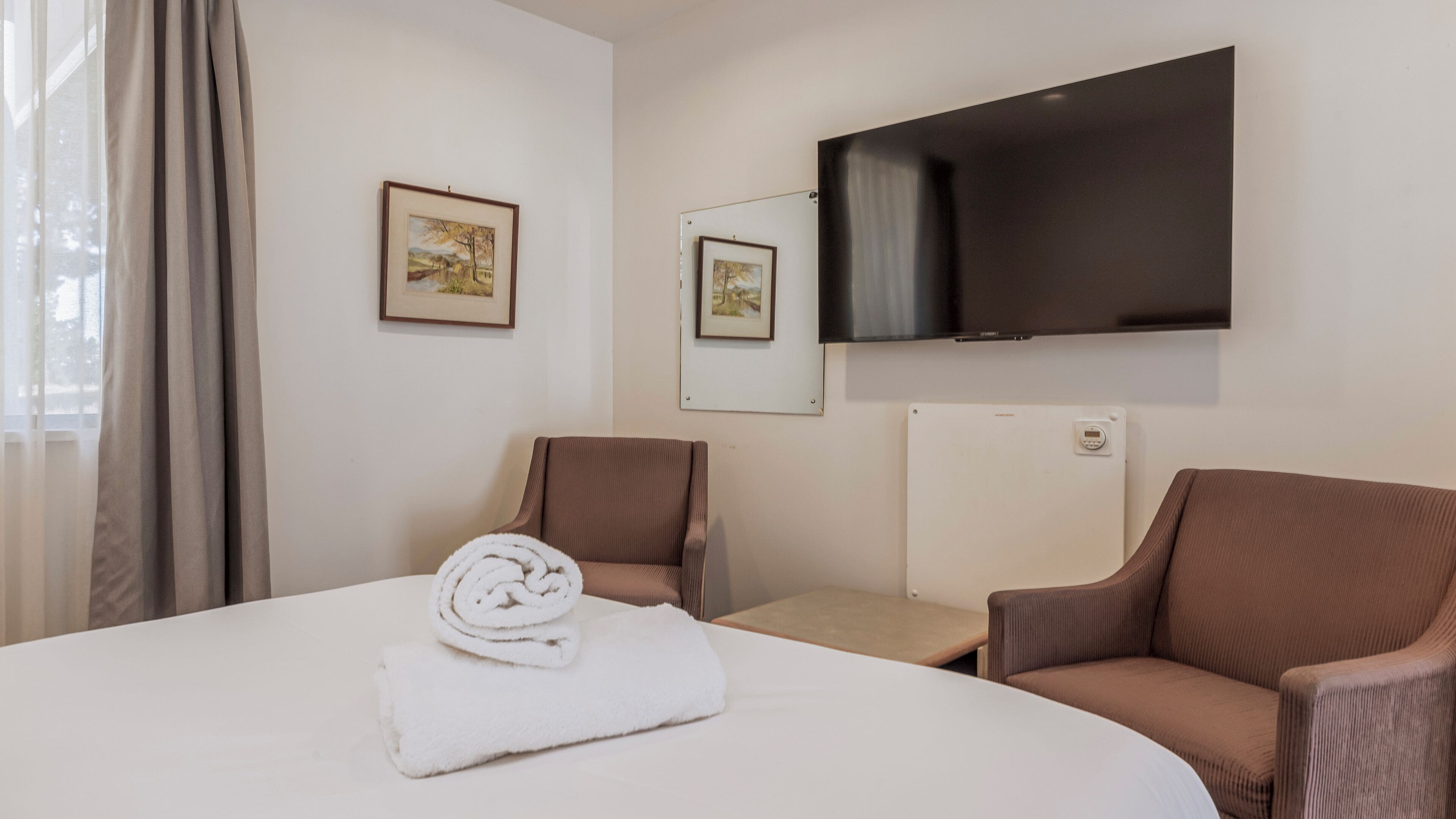 A hotel room with a bed, two brown armchairs, a TV mounted on the wall, and two framed pictures.