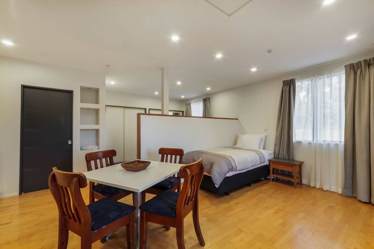 Ein Schlafzimmer mit Bett, Nachttisch, einem Fenster mit Vorhängen, Holzboden und einem Essbereich mit Tisch und Stühlen.