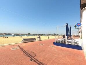 Un patio frente a la playa con mesas y sombrillas, con vista a una playa arenosa y aguas tranquilas.