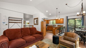 A modern living room with a red sofa, a wooden coffee table, and a brown armchair. A kitchen with wooden cabinets and a dining area with a table and chairs is visible in the background.