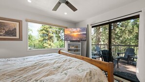 A bedroom with a bed, a ceiling fan, a TV, a window with a view of trees, and a balcony with chairs.