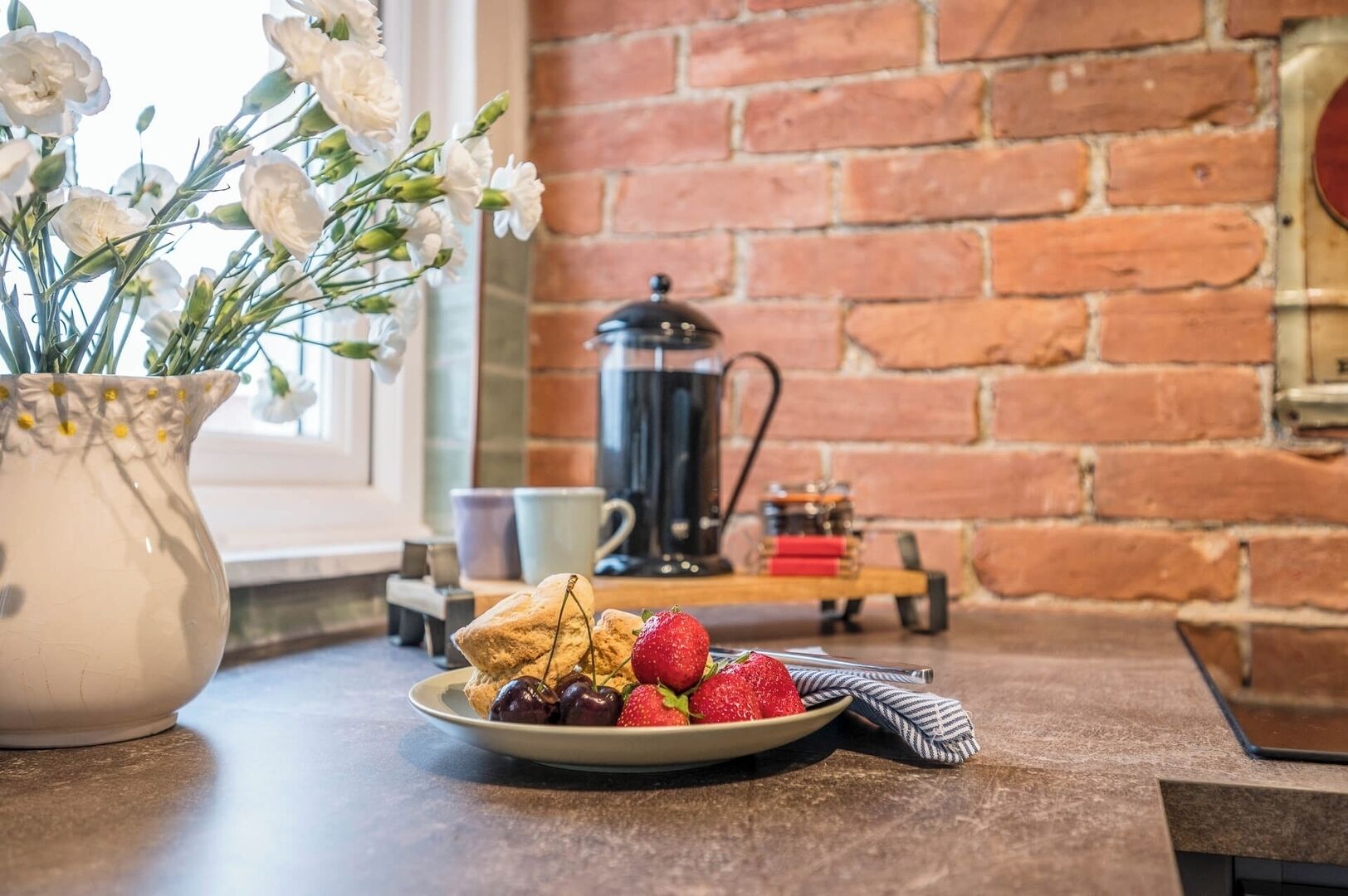A breakfast setting with a plate of strawberries and biscuits, a vase of white flowers, and a coffee pot on a brick wall background.