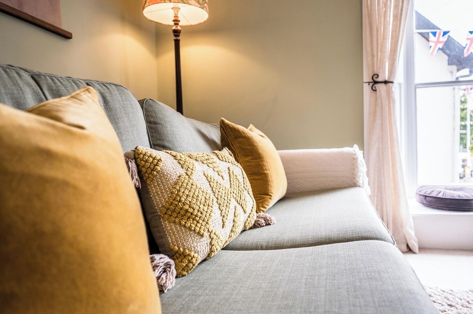 A cozy living room with a grey sofa, multiple cushions, a floor lamp, and a view of the outside through a window.
