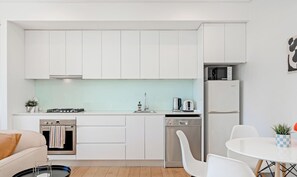 A modern kitchen with white cabinets, a stove, a dishwasher, and a refrigerator. There is a dining area with a table and chairs. - KozyGuru | Central Station | Stylish Urban Retreat (Surry Hills)