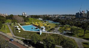 A large swimming pool with multiple lanes, surrounded by green lawns and palm trees, with a cityscape in the background. - KozyGuru | Central Station | Stylish Urban Retreat (Surry Hills)