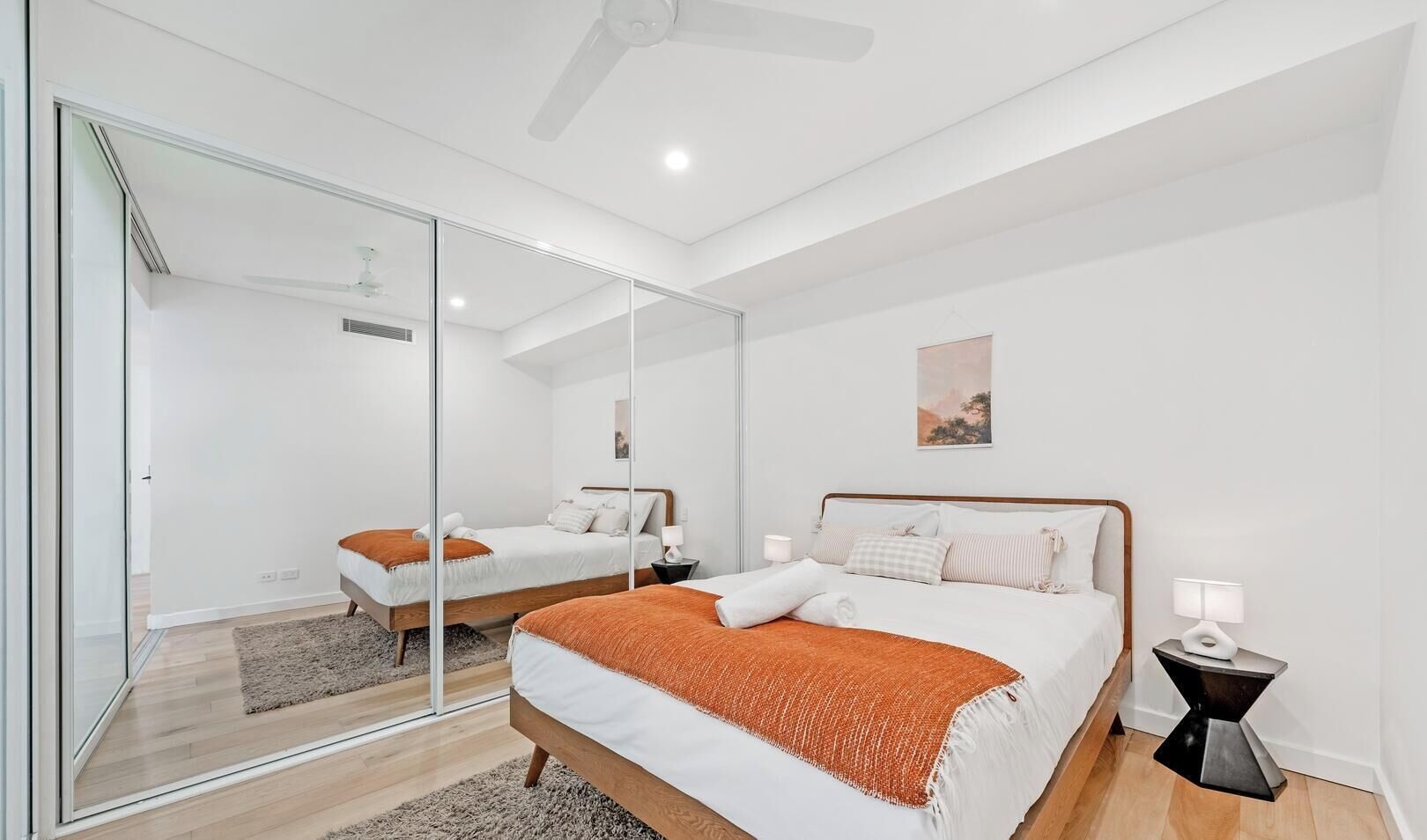 A modern bedroom with a large mirror, two beds, a ceiling fan, and a small bedside table with a lamp.