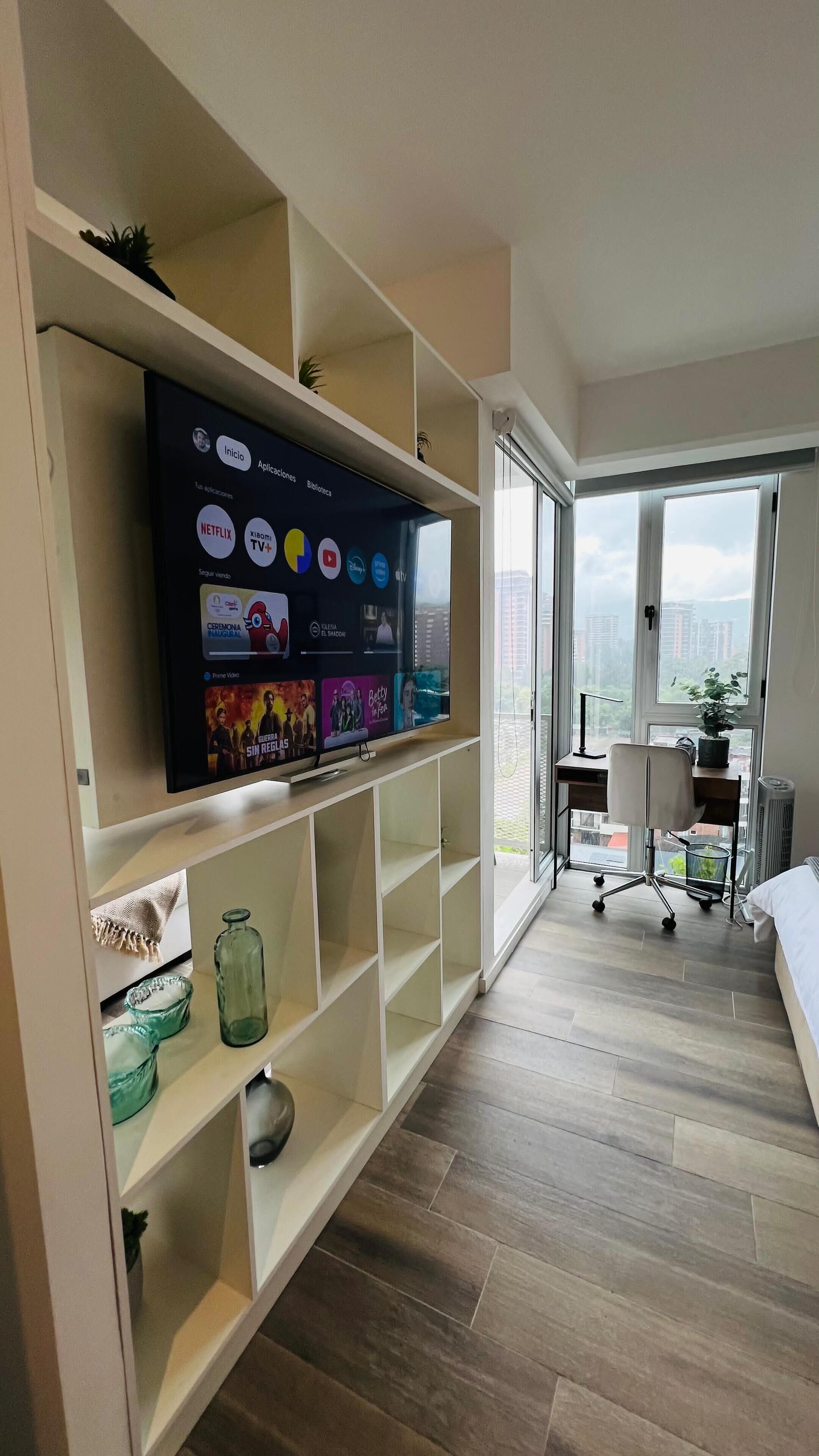 Un salon moderne doté d’un grand téléviseur à écran plat fixé au mur, d’un parquet et offrant une vue sur le paysage urbain grâce à une grande fenêtre.