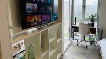 Un salon moderne doté d’un grand téléviseur à écran plat fixé au mur, d’un parquet et offrant une vue sur le paysage urbain grâce à une grande fenêtre.