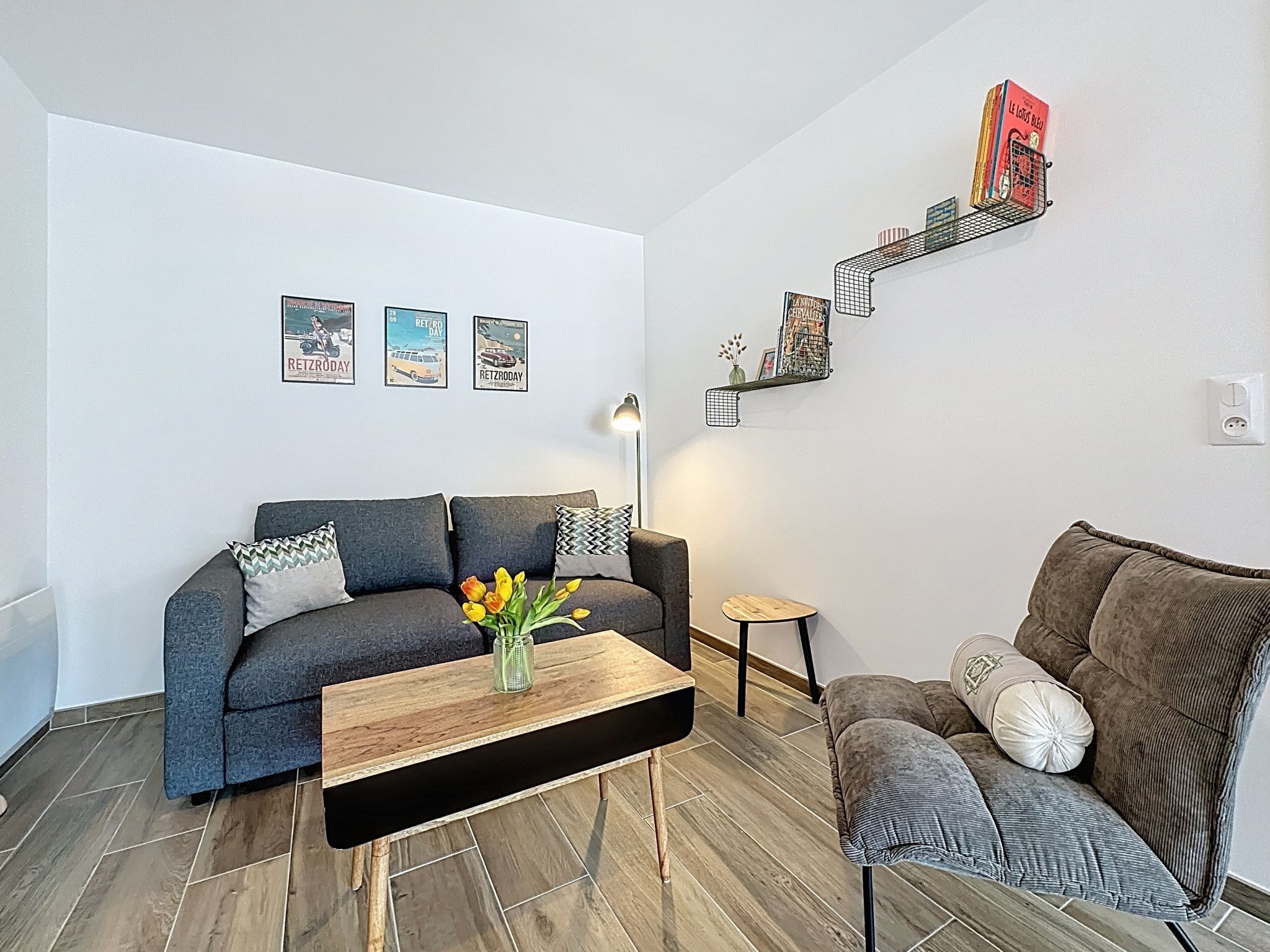 A modern living room with a grey sofa, a wooden coffee table with a vase of flowers, a small side table, and a bookshelf with books and decorative items.