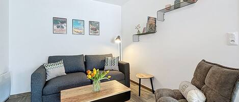 A modern living room with a grey sofa, a wooden coffee table with a vase of flowers, a small side table, and a bookshelf with books and decorative items.