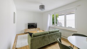 Uma sala de estar moderna com uma televisão de tela plana, um sofá verde, uma mesa branca e uma janela com vista para árvores.