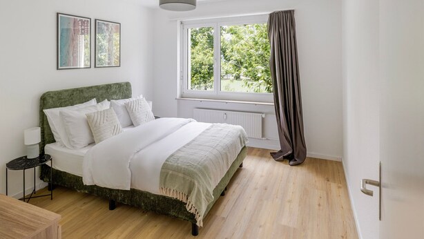 Quarto com cama, mesa de cabeceira, janela com cortinas e radiador.