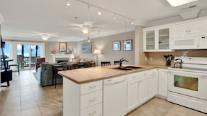 A spacious kitchen with white cabinetry, a large island, and a built-in oven. There is a dining area with a table and chairs, and a view of the outdoors through large windows. - Your Tybee Island Getaway! | Heated Pool Access (Tybee Island)
