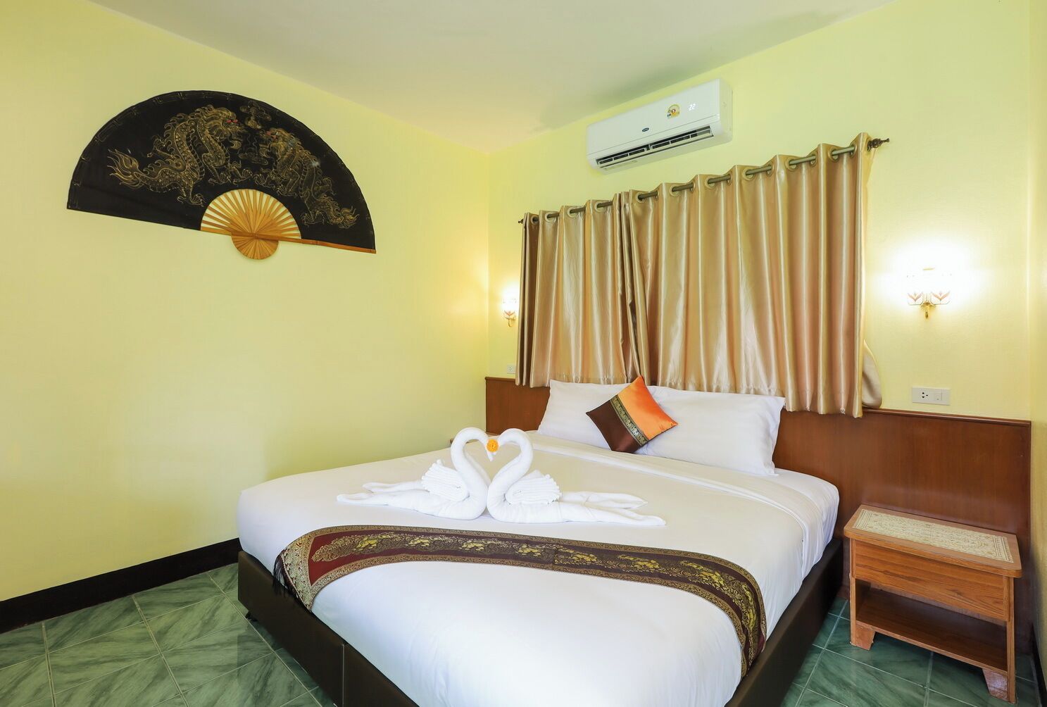 A hotel room with a bed, a wall-mounted air conditioner, a decorative fan, and a small wooden nightstand.