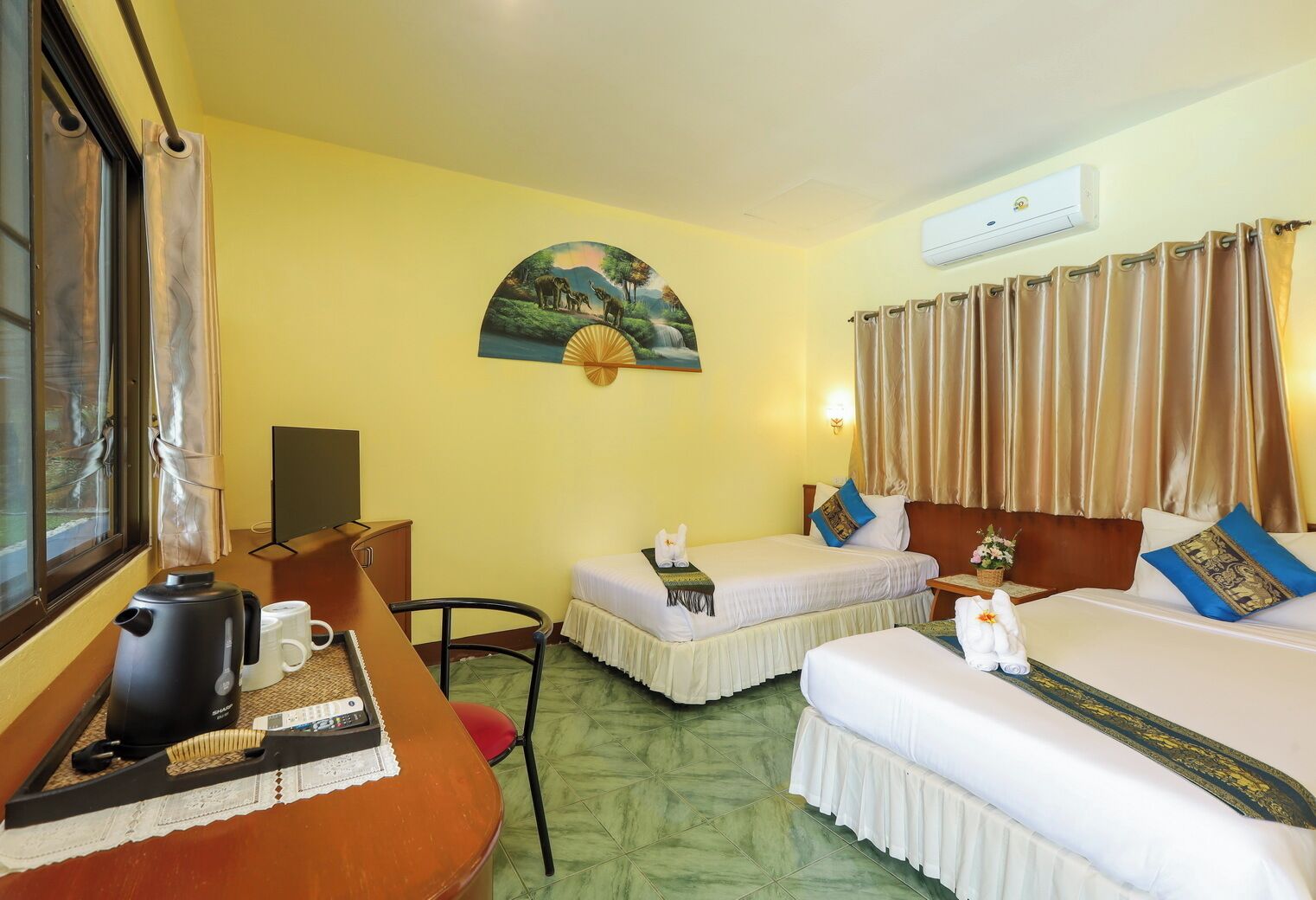 A hotel room with two beds, a desk with a kettle, a chair, a painting on the wall, and an air conditioning unit.
