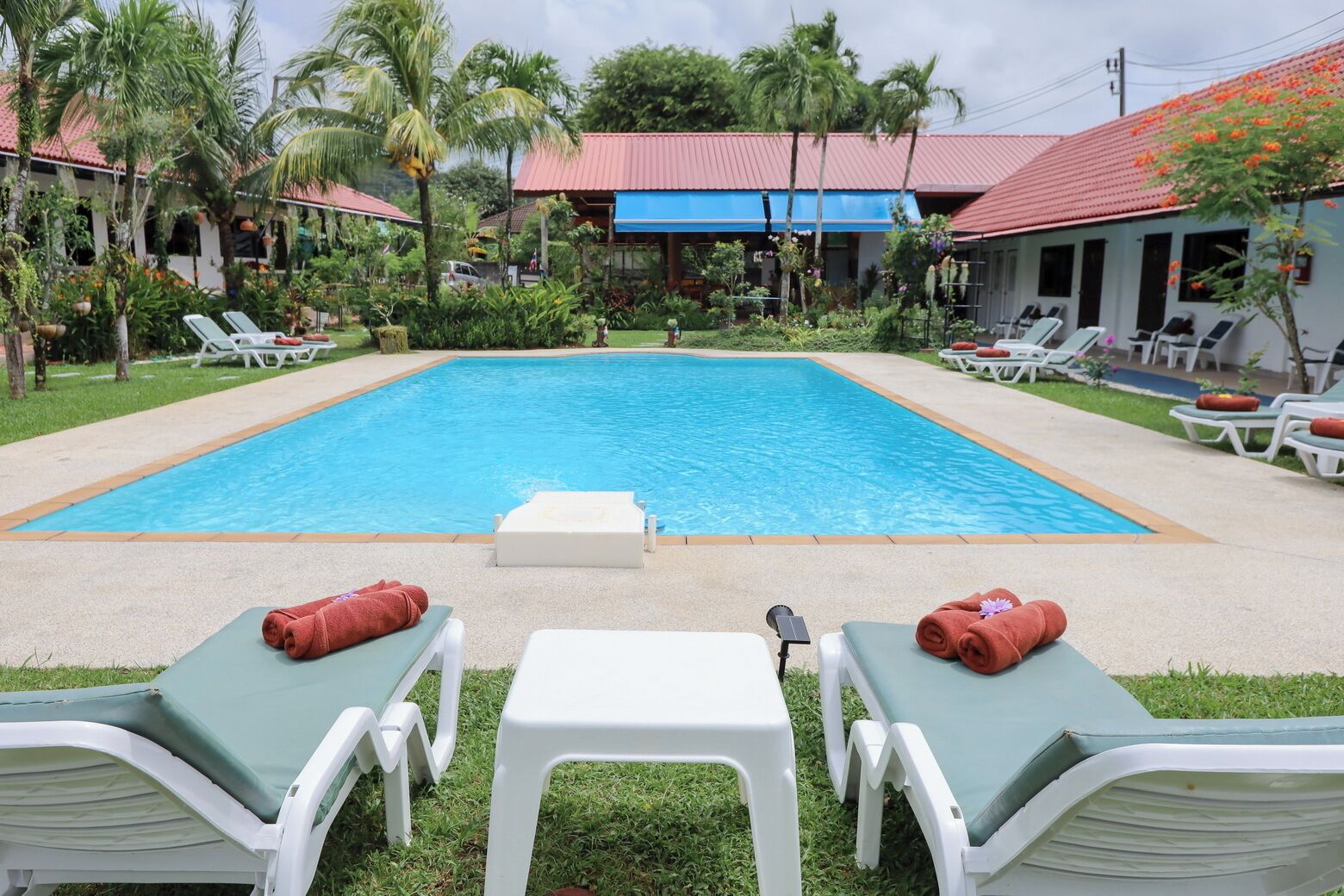 A swimming pool with lounge chairs and a small table in the center, surrounded by greenery and a building with a red roof.