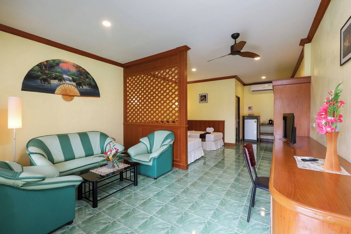 A hotel room with a green sofa set, a small table with a vase of flowers, a bed with two white bedsheets, a wooden partition, a ceiling fan, and a wooden reception desk.