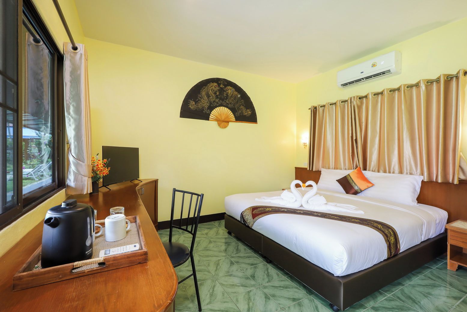 A hotel room with a bed, a desk with a kettle and cups, a chair, a fan, and a window with curtains.