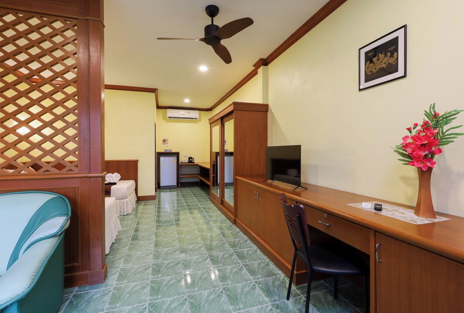 A hotel room with a bed, a desk, a chair, a television, a small refrigerator, and a decorative vase with flowers.