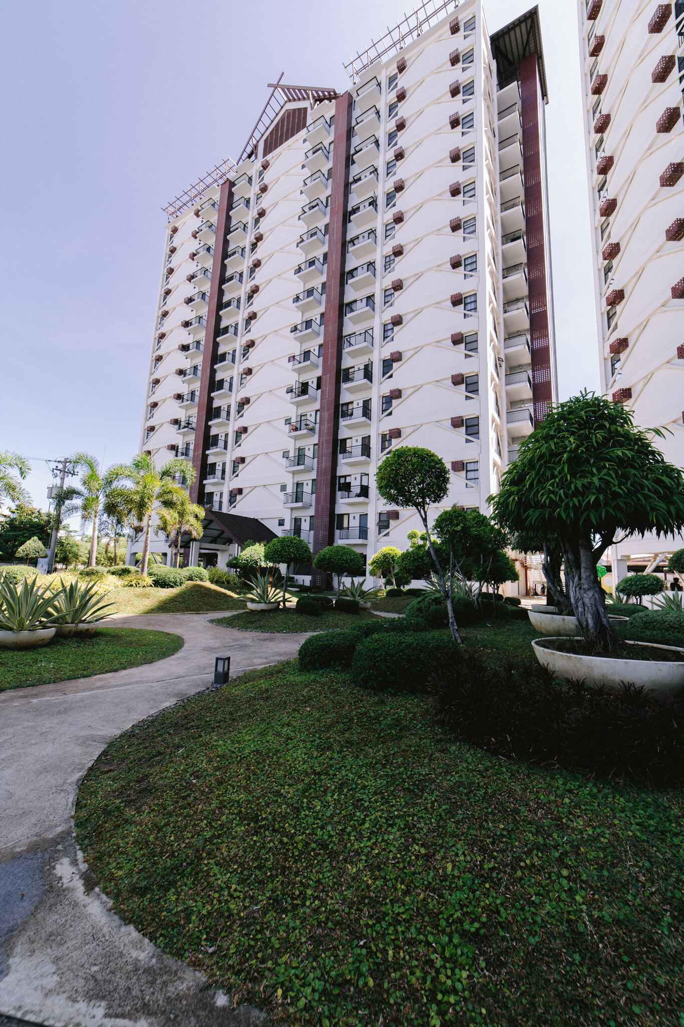 A high-rise residential building with multiple balconies, surrounded by well-maintained greenery and a paved pathway.