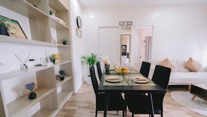 A dining area with a table set for four, surrounded by black chairs. There are shelves with various items and a potted plant on the left. A white sofa with beige cushions is in the background. - APARTMENT IN LAPU-LAPU CITY RESORT TYPE-Near Mactan Airport 60 SQM.WITH 2BR (Lapu-Lapu)