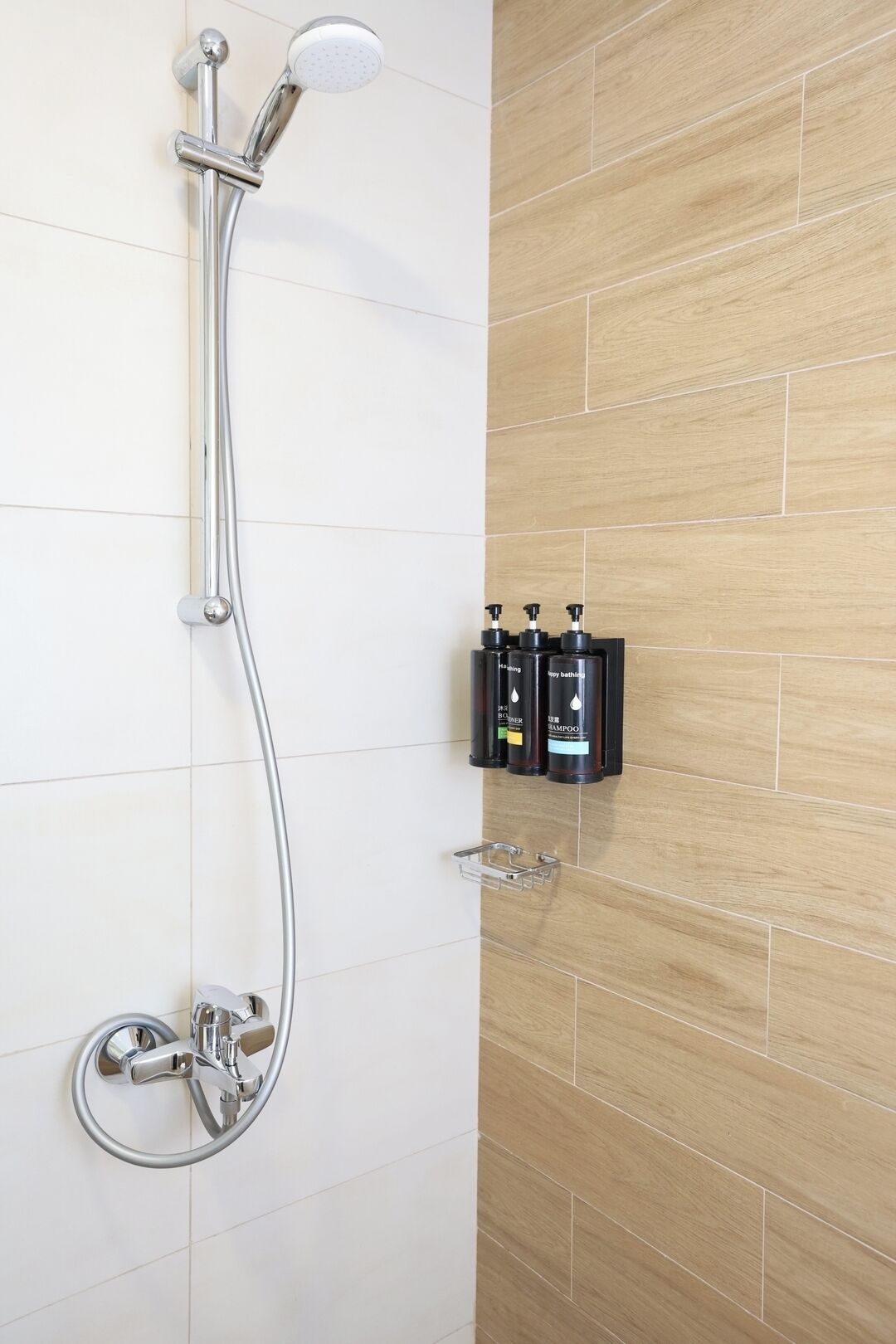 Une douche équipée d’un pommeau de douche à main et d’un robinet mural. Deux bouteilles de shampoing sont posées sur une étagère.