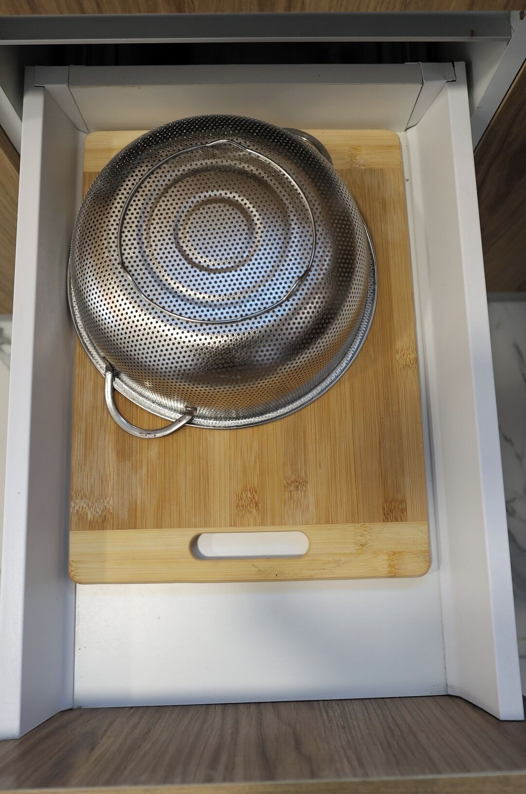 Une cuisine avec une planche à découper en bois et une passoire en métal à l’intérieur d’un meuble blanc.