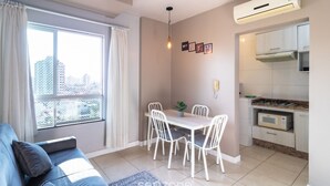 A modern living room with a sofa, dining table, and chairs. There is a window with curtains, an air conditioning unit, and a kitchen area with cabinets and a microwave. - Apto 1 Quarto no centro em Itajaí CST0905 (Itajai)