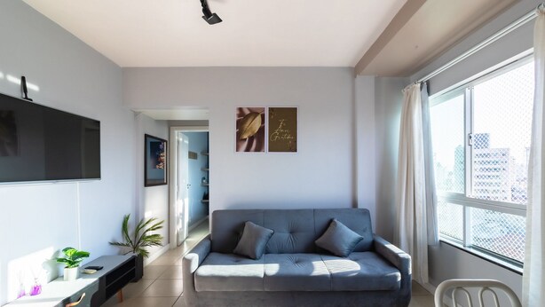 A modern living room with a grey sofa, a flat-screen TV mounted on the wall, and a large window with a view of buildings. - Apto 1 Quarto no centro em Itajaí CST0905 (Itajai)
